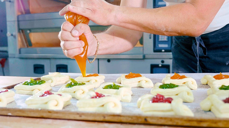 Close-up of a baker piping jam onto uncooked pastries in a professional kitchen setting