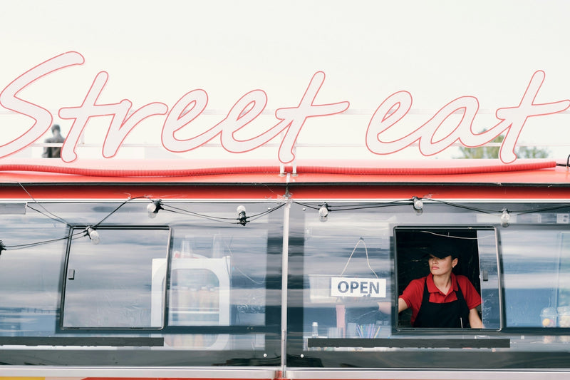 Female vendor standing at the open window of a food truck under a neon "Street Eat" sign