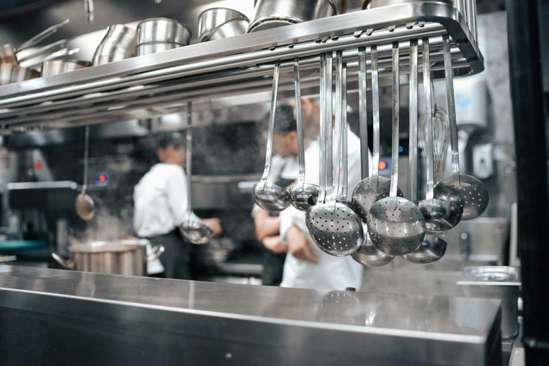 Blurred commercial kitchen background with chef and stainless steel utensils in foreground