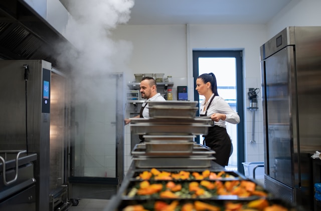 Two chefs working in a commercial kitchen with steaming ovens and trays of food
