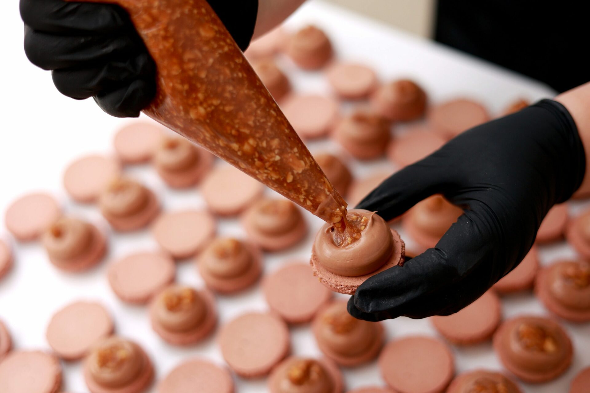 Gloved chef piping chocolate filling into macarons in a professional kitchen.