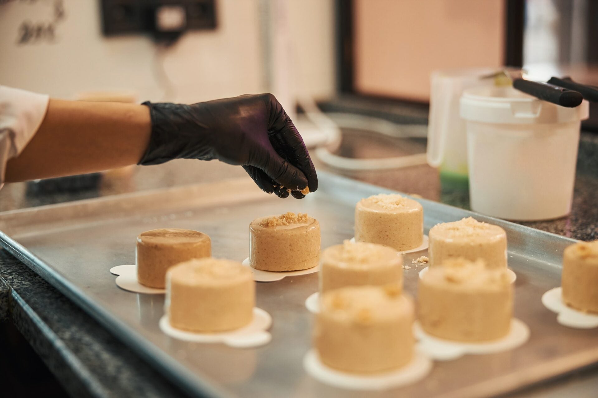 Chef with gloved hand sprinkling crumble topping onto mini cakes on a baking tray