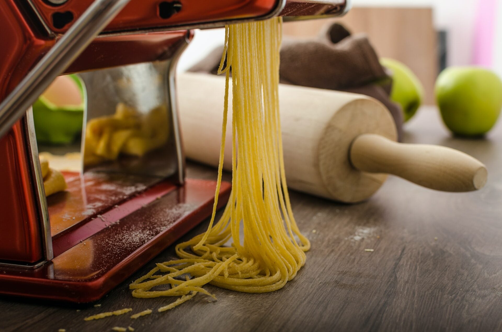 Fresh pasta strands being extruded from a red pasta machine, with a wooden rolling pin in the background